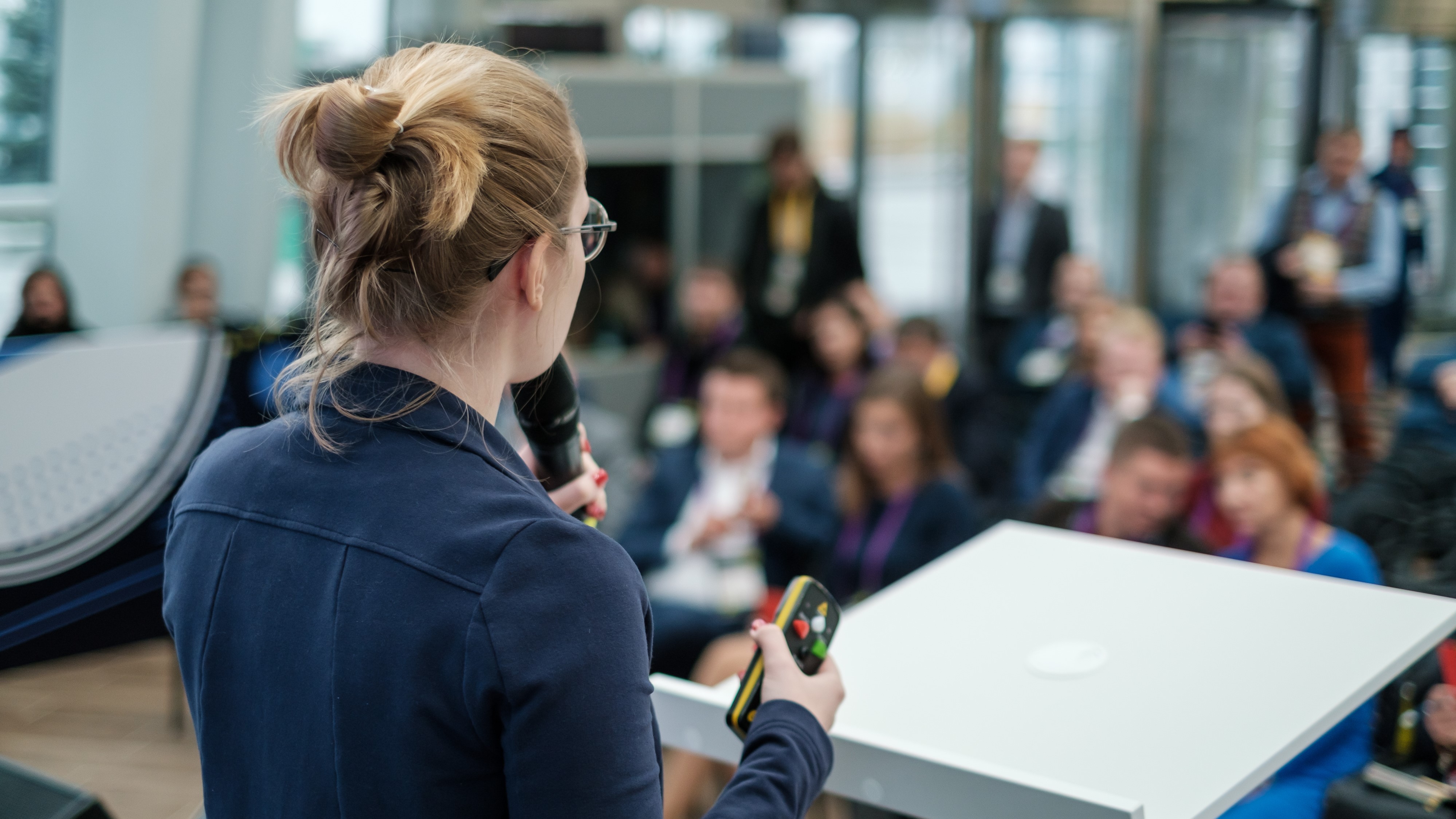 Instructor stands at a podium facing the class while holder a clicker in one hand, and speaking into a microphone.